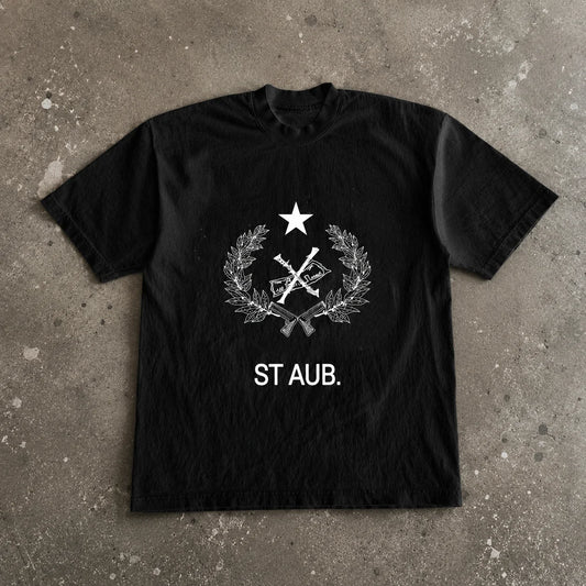 Modèle portant l'OG Saint Aub Black T-shirt. Noir pur, sérigraphie blanche emblème héritage central. Contexte urbain brut — béton, rue authentique. Coton premium 220g/m², coupe oversize pensée pour durer partout. C'est l'original. L'uniform intemporel de ceux qui restent vrais. Édition limitée. Pas de restock.